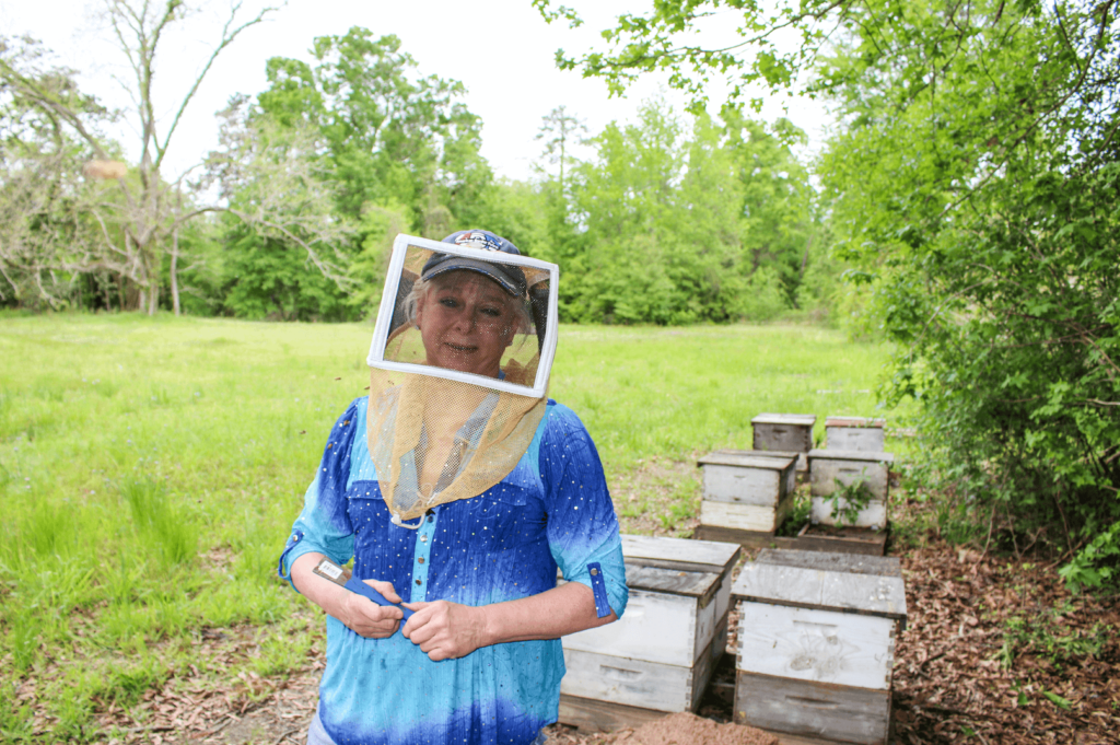 Photo of Beekeeper Jane Collins in one of her bee yards. She is wearing a beekeeper veil and standing next to beehives.