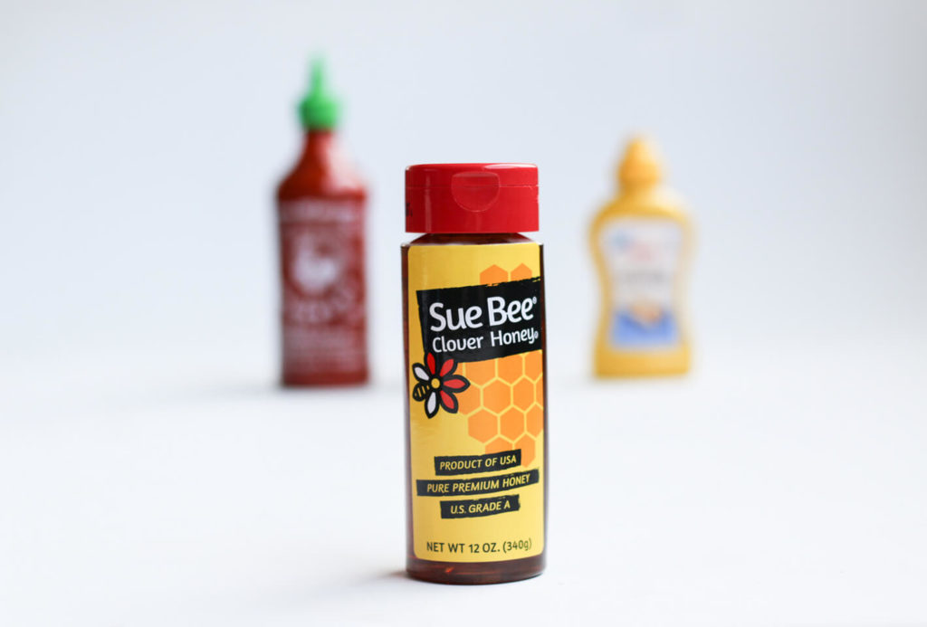 Picture of a bottle of Sue Bee Honey with Sriracha and mustard in the background. 